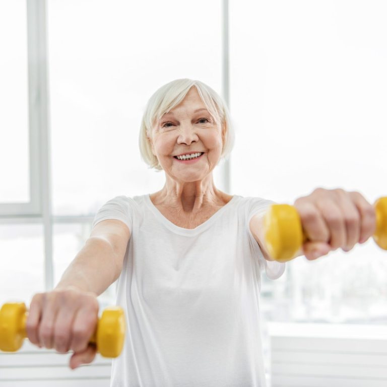 Sport is my life. Cheerful mature woman is training with yellow dumbbells. She is looking at camera and smiling
